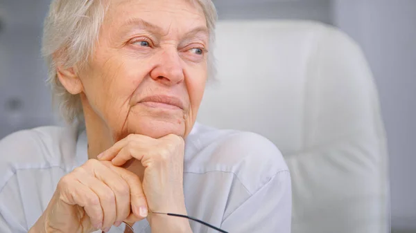 Femme âgée pensionnée tient la tête sur les mains ridées avec des lunettes et regarde de côté avec le sourire assis sur la chaise à la maison — Photo