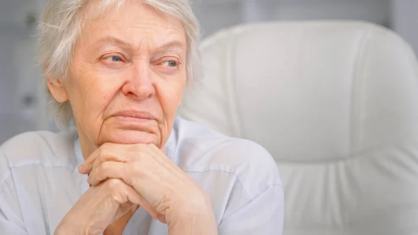 Bedachtzame grijze harige senior dame kijkt in het raam houden hoofd over handen en zitten op witte stoel in het kantoor — Stockfoto