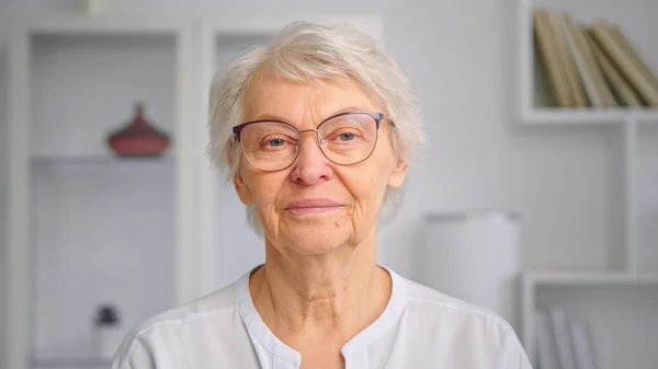 Aged lady with short grey hair and glasses looks straight and smiles cheerfully standing against white furniture — Stock Photo, Image