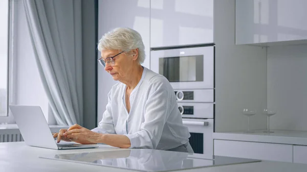 Empresária idosa concentrada com cabelo cinza curto e tipos de óculos no laptop sentado na cozinha em casa — Fotografia de Stock