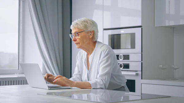 Old Woman Company Manager types on grey laptop κάθεται σε μεγάλο λευκό τραπέζι στην κουζίνα κατά παράθυρο με κουρτίνες — Φωτογραφία Αρχείου