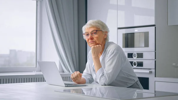 Deprimerad pensionär affärskvinna typer på grå laptop sitter i köket — Stockfoto