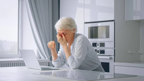 Depressed tired of work lady pensioner with short grey hair massages temples with hands sitting by laptop — Stock Photo, Image