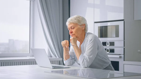 Thoughtful senior lady secretary with short hair holds glasses in hand and sits at grey laptop in kitchen — Stock Photo, Image