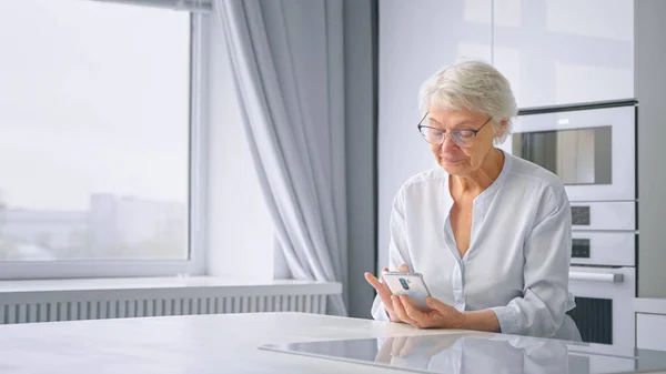 Smiling pensioner lady in glasses waves hand and talks at online video call holding grey smartphone sitting in kitchen — Stock Photo, Image