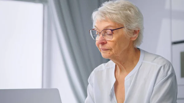 Upset aged businesswoman with grey hairstyle wipes nose with paper napkin and types on laptop sitting at home — Stock Photo, Image