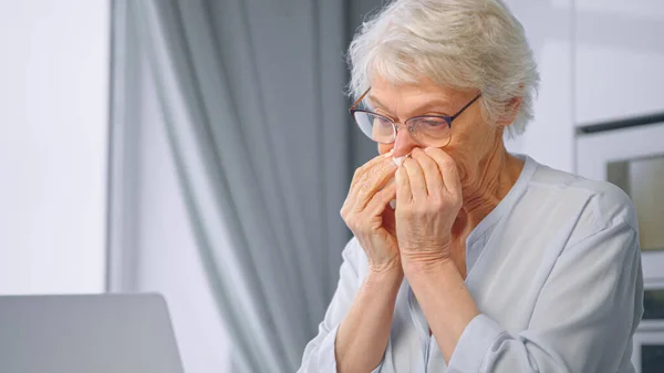 Ill aged secretary with grey hair wipes red nose with white paper napkin and types on laptop at home isolation — Stock Photo, Image