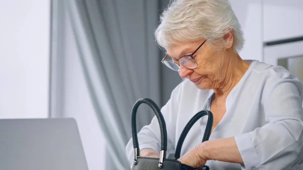 Aged grey haired businesswoman rummages in black leather handbag and returns to work typing on laptop sitting at home — Stock Photo, Image