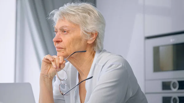 Nachdenkliche Seniorin mit kurzen Haaren hält Brille in der Hand und sitzt zu Hause am grauen Laptop in der Küche — Stockfoto