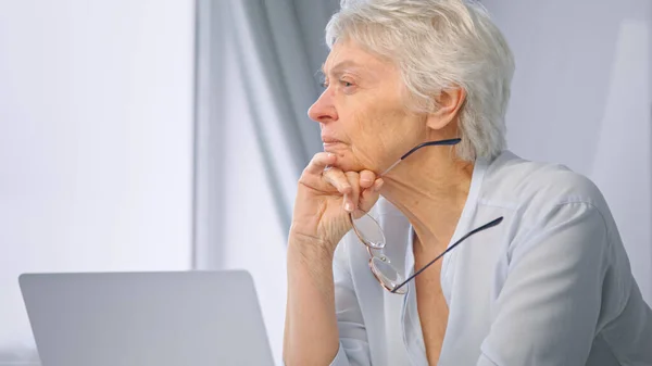 Thoughtful senior lady secretary with short hair holds glasses in hand and sits at grey laptop — Stock Photo, Image