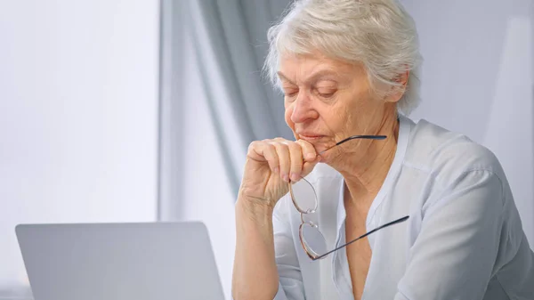 Ragionevole segretaria anziana con i capelli corti tiene gli occhiali in mano e si siede al computer portatile grigio in cucina a casa — Foto Stock