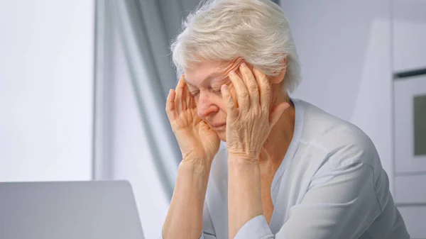 Уставшая от работы женщина-пенсионер с короткими седыми волосами массирует виски с руками, сидя на ноутбуке вблизи — стоковое фото
