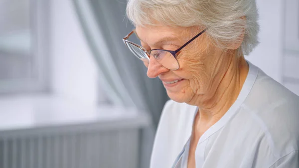 Mujer mayor encantada con pelo gris corto y tipos de gafas en el teléfono inteligente blanco y sonrisas contra la ventana del hogar — Foto de Stock