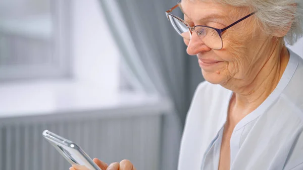 Abuela de pelo gris en gafas mira hacia abajo y sonríe alegremente sentado contra la ventana borrosa en casa — Foto de Stock