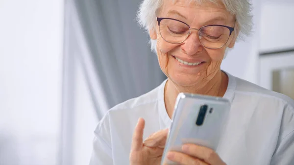 Happy aged in glasses and blue blouse communicates with children on white smartphone against blurred window at home — Stock Photo, Image