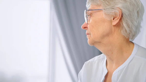 Ragionevole donna anziana con capelli corti grigi e occhiali guarda fuori sfocato grande finestra con luce seduta a casa — Foto Stock