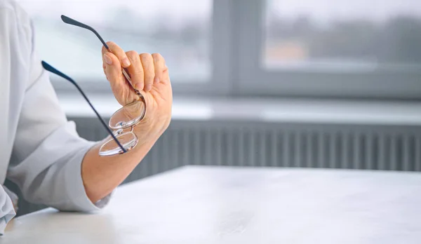 Tänksam gammal affärskvinna i blå blus hand håller glasögon över vitt bord mot suddig stora hem fönster — Stockfoto