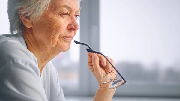 Vecchia nonna tiene gli occhiali in mano rugosa con il viso serio seduto contro la finestra sfocata sotto la luce del sole a casa — Foto Stock