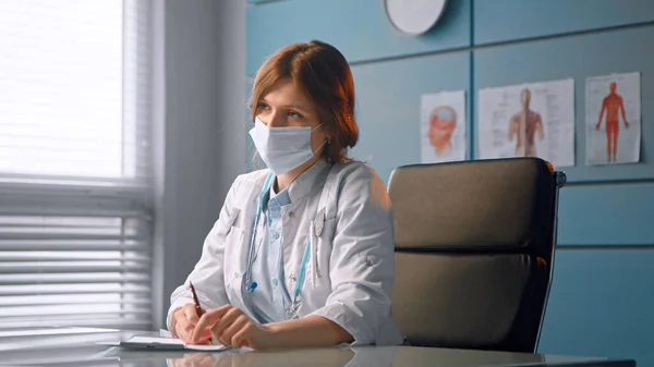 Молодая женщина врач в белом медицинском халате разговаривает с новым пациентом и пишет историю болезни перед обследованием — стоковое фото