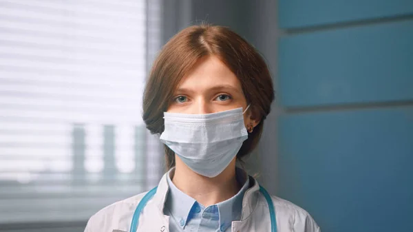 Mujer pelirroja con máscara facial y se ve recta de pie en la habitación del hospital local — Foto de Stock