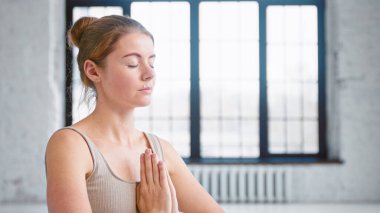 Bej rengi elbiseli sarışın kadın hafif yoga stüdyosunda namaste Mudra 'da el ele tutuşarak derin bir nefes alıyor.