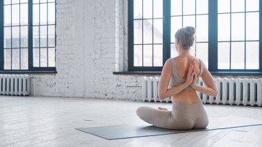 Eşofmanlı, saçları topuz gibi olan zarif sarışın yoga uygulayıcısı stüdyonun penceresinin yanında, yerde ters namaste yapıyor.