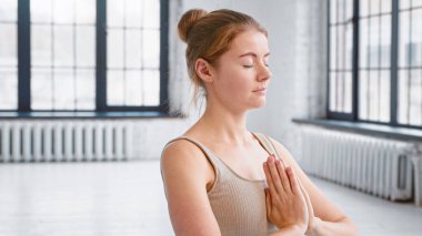 Konsantre olmuş, rahat eşofmanlı genç bir kadın geniş yoga stüdyosunda namaste Mudra ile meditasyon yapıyor.