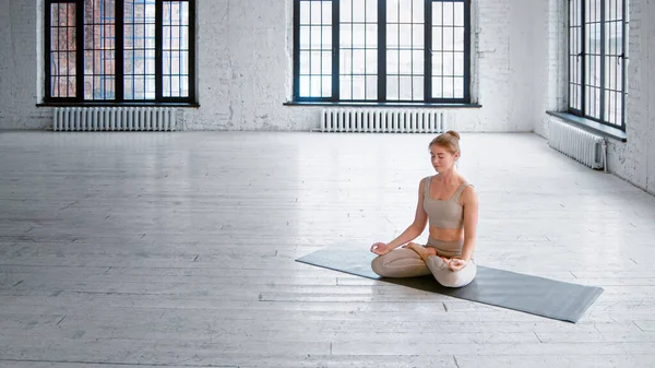 Mujer rubia sonriente en chándal elegante se sienta en la estera en postura de loto meditando en un estudio amplio y luminoso —  Fotos de Stock