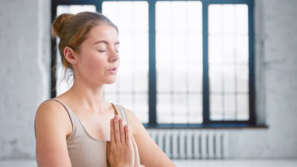 Mujer joven y tranquila con moño de pelo en top beige hace ejercicios de respiración de yoga tomados de la mano en namaste mudra en estudio —  Fotos de Stock