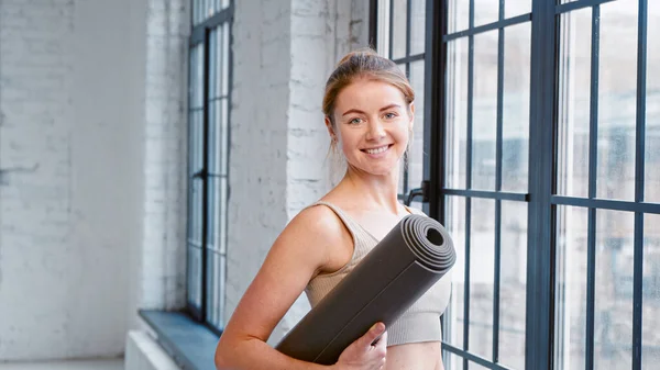 Entraîneur de fitness femme blonde avec un chignon de cheveux tenant tapis regarde par la fenêtre et se tourne vers la caméra dans le studio de yoga moderne — Photo