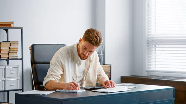 Bearded accountant sums up unexpected expenses from checks on calculator and writes results in paper notebook — Stock Photo, Image