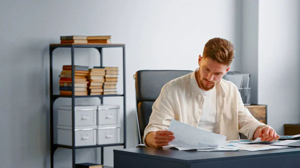Skilled bearded accountant looks through reports on sheets of papers sitting at table with calculator in bank office — Stock Photo, Image