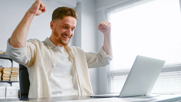 Концентрированный бизнесмен подписывает контракт онлайн и поднимает руки со счастьем сидя рядом с белым ноутбуком — стоковое фото