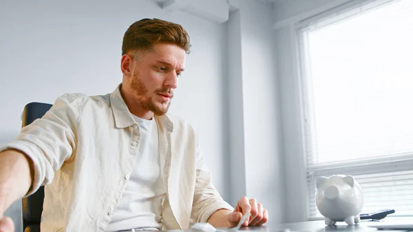 Baard bedrijf werknemer in geel shirt schudt witte spaarvarken bank om munten te luisteren knipperen over kantoor tafel — Stockfoto