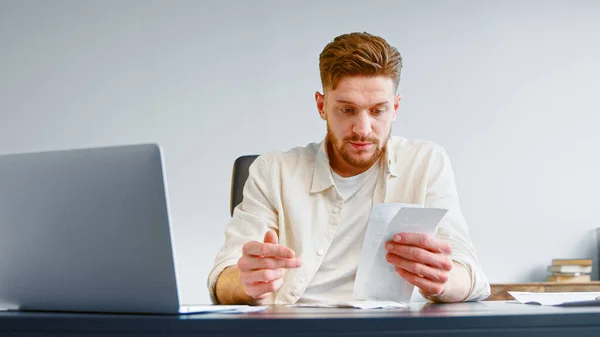 Бородатый профессиональный бухгалтер сортирует бумажные квитанции счета, сидя за столом с белым ноутбуком — стоковое фото