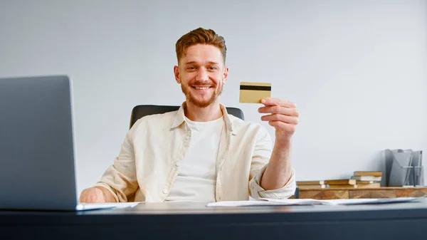 Успешный менеджер компании с бородой держит кредитную карту и позирует сидя за серым столом с ноутбуком и бумагами — стоковое фото