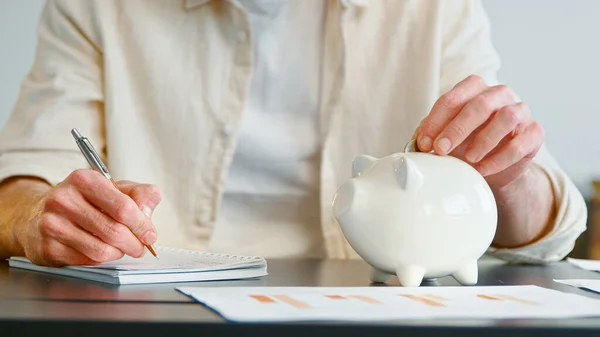 Oigenkännlig man husdjur vit spargris bank och skriver rapport i papper anteckningsbok — Stockfoto