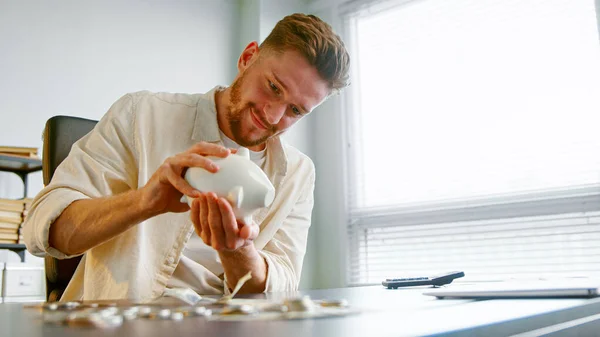 Allegro ragazzo manager con barba scuote salvadanaio bianco per prendere l'ultima moneta sopra tavolo grigio Fotografia Stock