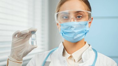 Mavi tek kullanımlık gözlüklü ve beyaz önlüklü genç bir kadın şeffaf bir aşı şişesi taşıyor.