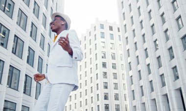 Turuncu tişörtlü ve takım elbiseli Afrikalı Amerikalı adam büyük ofis binaları arasında dans ediyor.