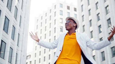 Kulaklıklı ve gözlüklü Afrikalı Amerikalı adam beyaz gökdelenler arasında kare üzerinde dans ediyor.