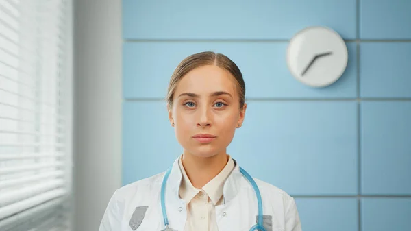 Blonde junge Allgemeinmedizinerin im weißen Mantel mit blauem Stethoskop sieht gerade aus — Stockfoto