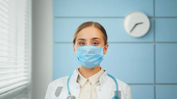Mujer joven médico general en abrigo blanco y máscara facial desechable azul se ve recto —  Fotos de Stock