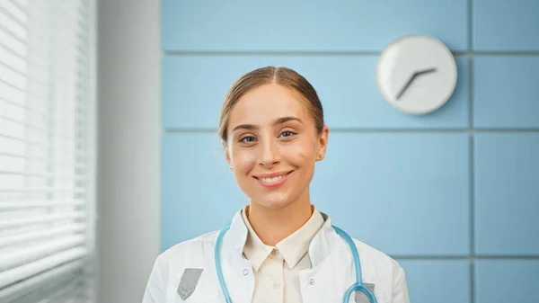 Blond ung kvinna allmänläkare i vit rock med blå stetoskop ser rakt stående — Stockfoto