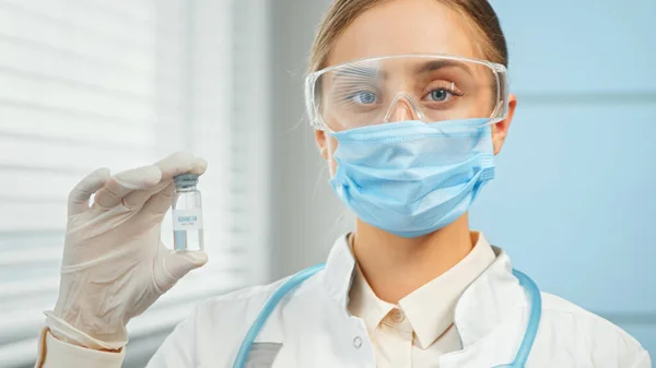Jovem mulher de óculos de máscara descartáveis azuis e casaco branco contém frasco transparente com vacina — Fotografia de Stock