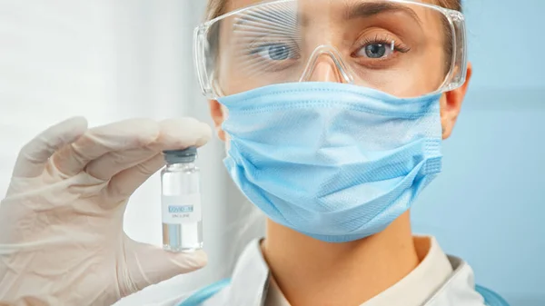 Lady investigadora en gafas de mascarilla desechables y guantes estériles sostiene el vial de vacuna en la mano —  Fotos de Stock