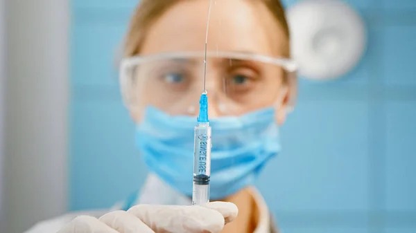 Enfermeira loira desfocada em azul máscara facial descartável e óculos pressiona seringa e pulveriza vacina — Fotografia de Stock