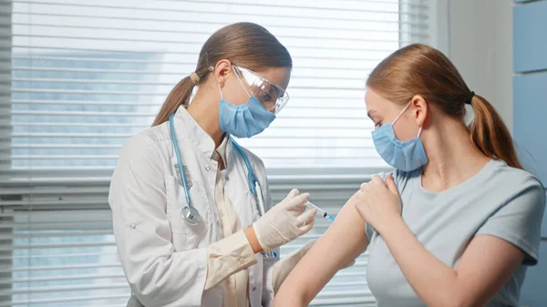 Blonde lady hospital nurse in disposable facial mask and glasses injects young woman patient arm — Stock Photo, Image