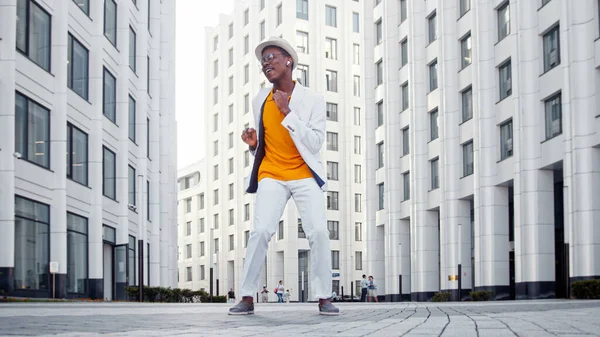 Black guy in orange t shirt under white jacket with hat and sunglasses dances on grey paved road — Stock Photo, Image