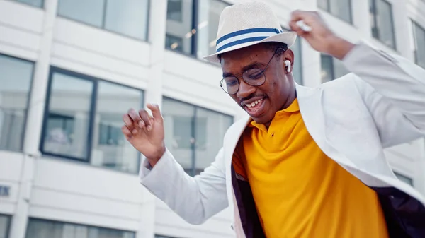 African American man in hat and wireless headphones listens to music and dances with joyful smile on megalopolis street — Stock Photo, Image